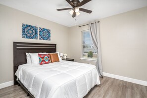 A bedroom with a bed, a ceiling fan, two wall art pieces, a window with curtains, and a nightstand with a lamp and flowers.