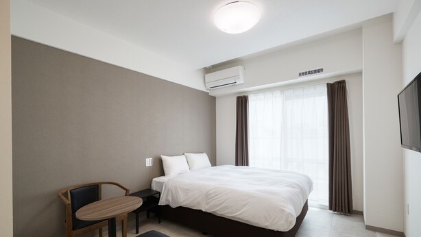 A hotel room with a bed, a desk, a chair, and an air conditioning unit.