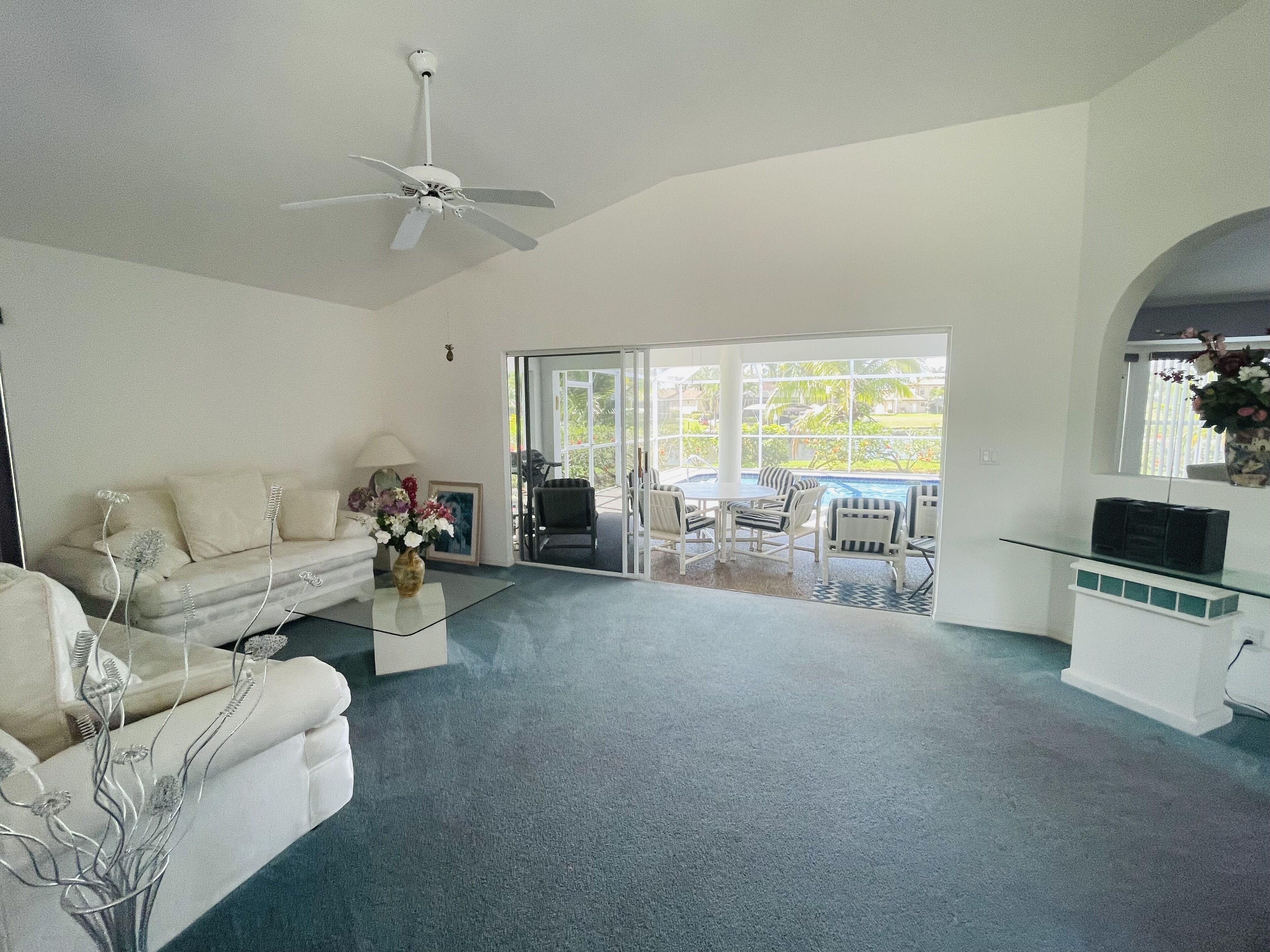 A spacious living room with a white sofa, a coffee table, and a TV on a glass cabinet. There is a ceiling fan and a view of an outdoor patio with a pool.