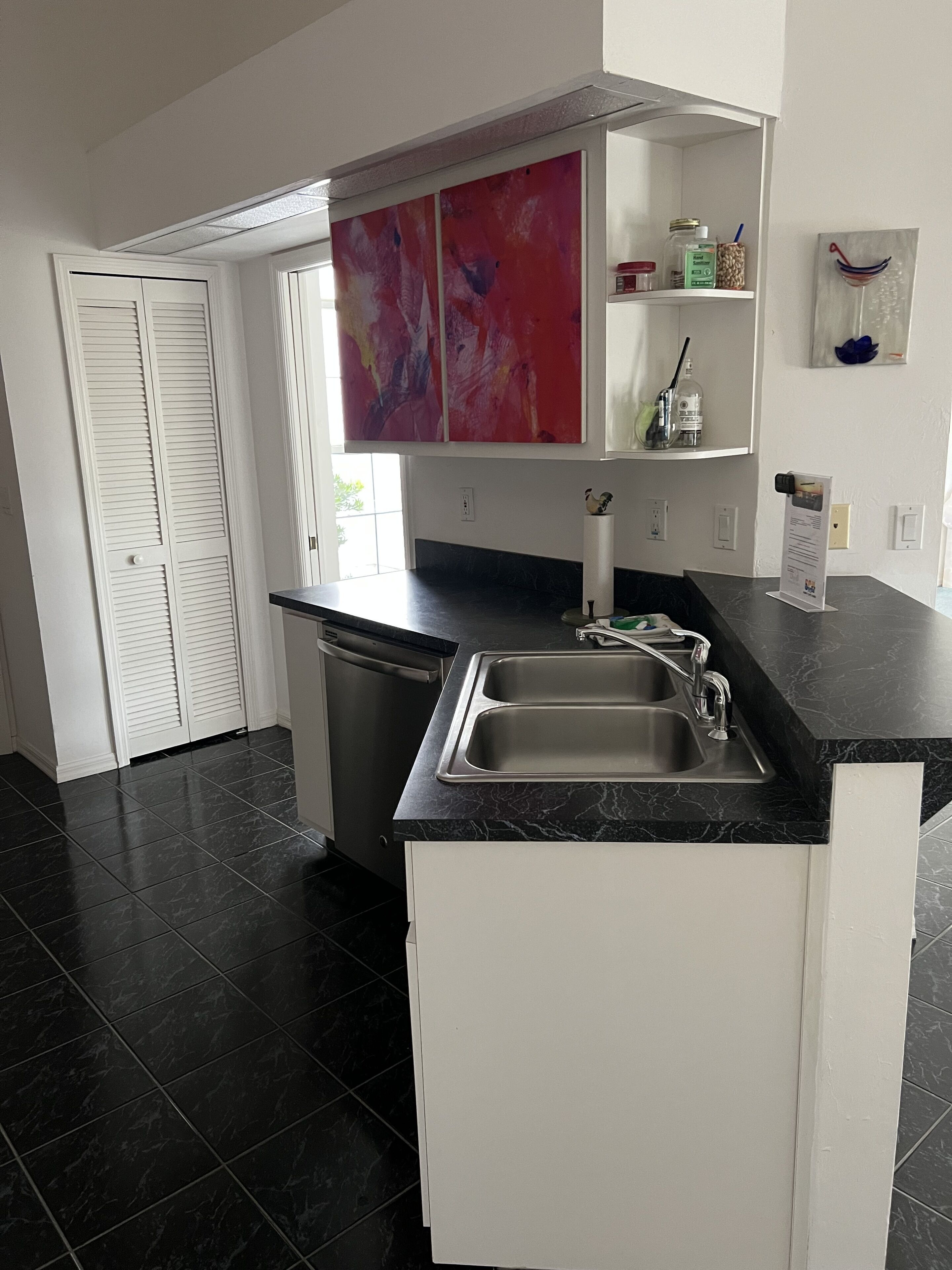 Cocina moderna con encimera de mármol negro, pileta de acero inoxidable y muebles blancos. Se observa una ventana con vista a áreas verdes, y un cuadro abstracto rojo sobre la encimera.