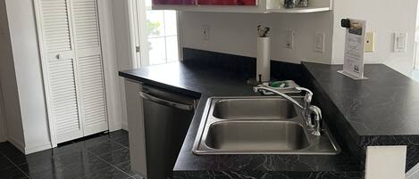 Cocina moderna con encimera de mármol negro, pileta de acero inoxidable y muebles blancos. Se observa una ventana con vista a áreas verdes, y un cuadro abstracto rojo sobre la encimera.