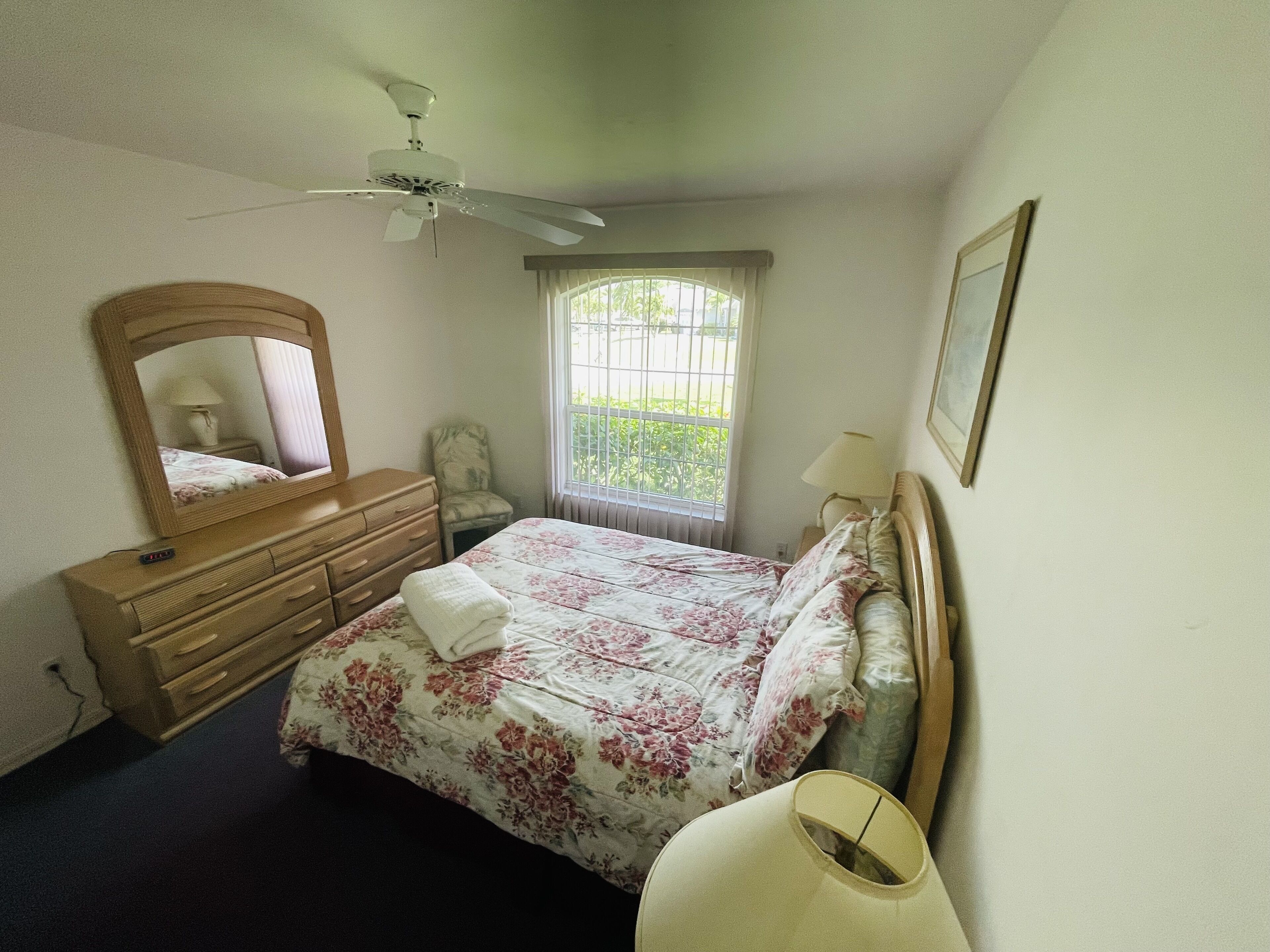 A bedroom with a bed, dresser, chair, window, ceiling fan, and lamp.