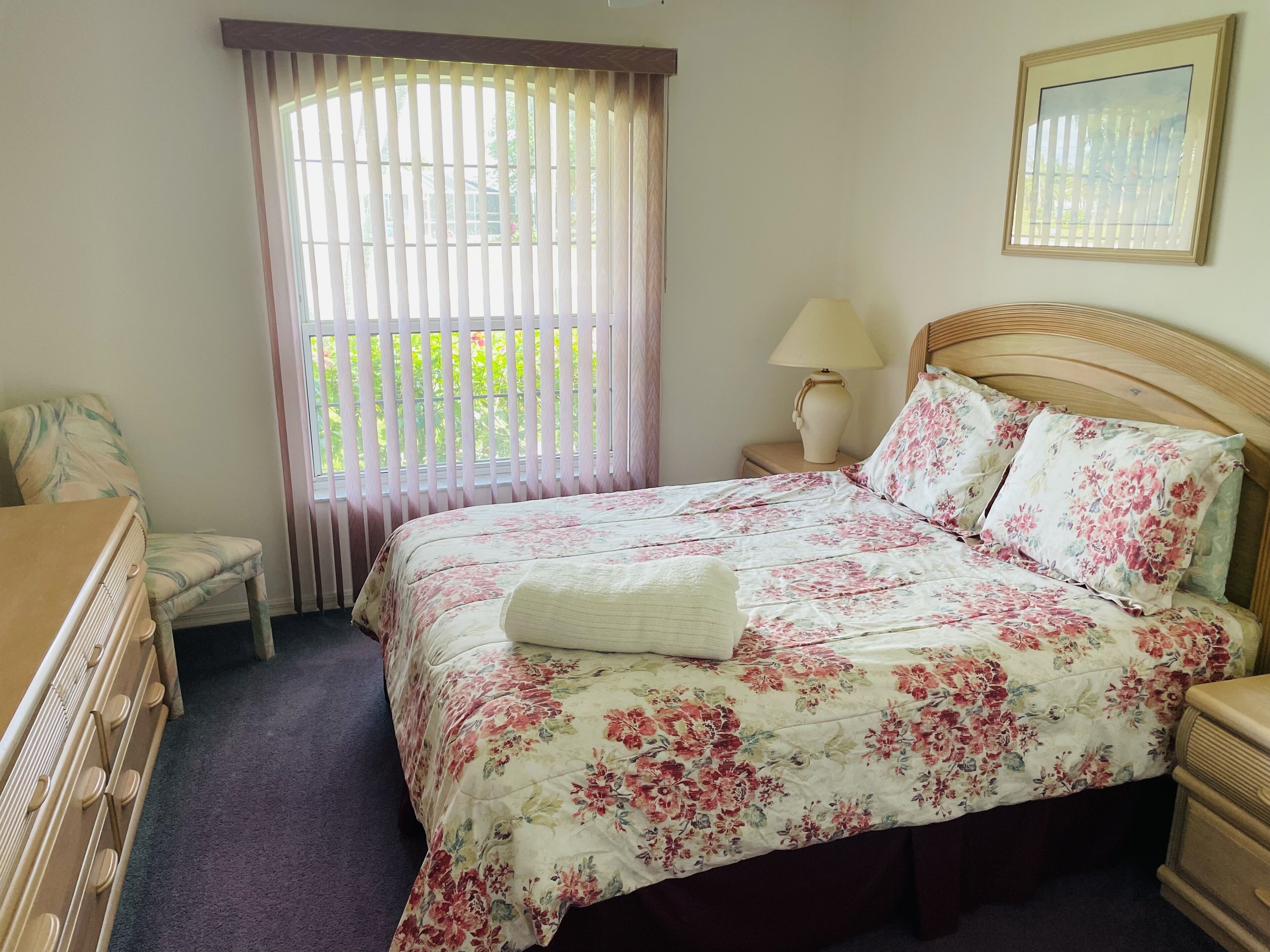 A bedroom with a floral bedspread, a wooden headboard, a bedside lamp, a chair, and a window with blinds.