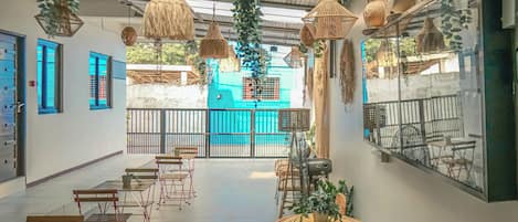An indoor space with tables and chairs, hanging wicker lamps, and a view of a blue building outside.