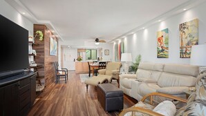 Un salon moderne avec un téléviseur à écran plat, un plancher en bois, un canapé, une table à manger et une cuisine en arrière-plan.