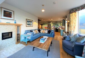 A living room with a fireplace, a blue sofa, a wooden coffee table, and a kitchen in the background.