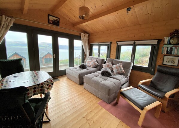 A cozy room with a wooden ceiling, a grey sofa with plaid cushions, a black leather chair, and a round table with a polka dot tablecloth.