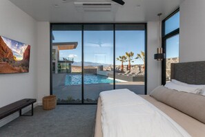 A modern bedroom with a large bed, a bench, a basket, and a view of a swimming pool and mountains. - Eleanor at Copper Rock Golf Course, Private Pool, 2 Hot Tubs, Basketball Court, and more (Hurricane)