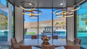 A conference room with a large table, six chairs, and a decorative bowl centerpiece. The room has large windows offering a view of a swimming pool and mountains. - Eleanor at Copper Rock Golf Course, Private Pool, 2 Hot Tubs, Basketball Court, and more (Hurricane)