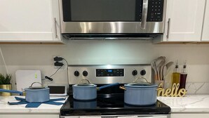 Et køkken med komfur i rustfrit stål, en mikrobølgeovn ovenover, og hvide skabe. Der står to blå gryder på komfuret, og der er et skilt med ordet 'hello' på disken.