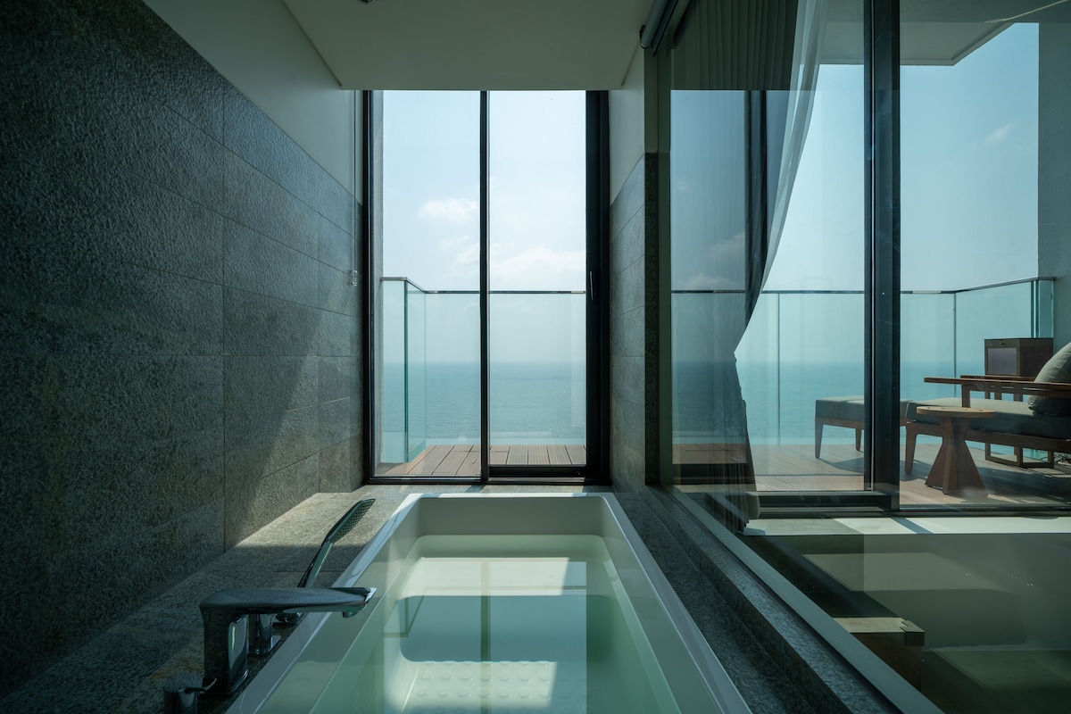Une salle de bain moderne avec un jacuzzi entouré de parois en verre, de grandes fenêtres et une vue sur la mer.