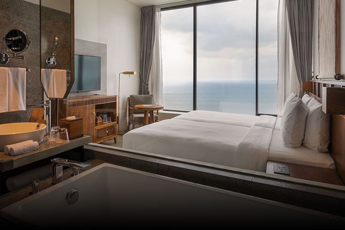 Habitación de hotel moderna con una bañera grande, un tocador con un espejo redondo, un televisor de pantalla plana y vista al océano.