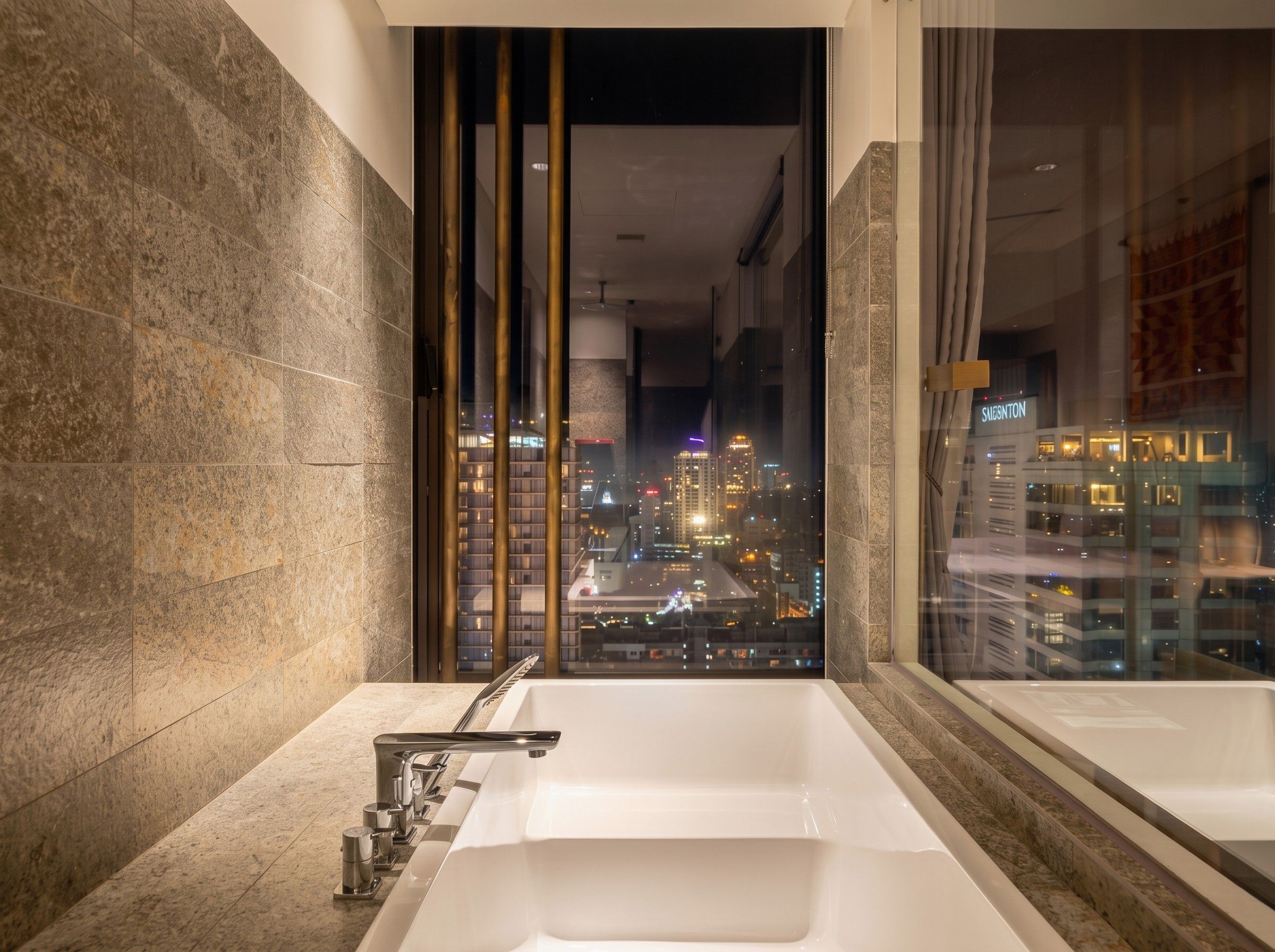A modern bathroom with a large bathtub, a city view through a glass door, and a stone wall.