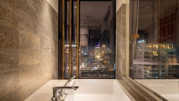 A modern bathroom with a large bathtub, a city view through a glass door, and a stone wall.