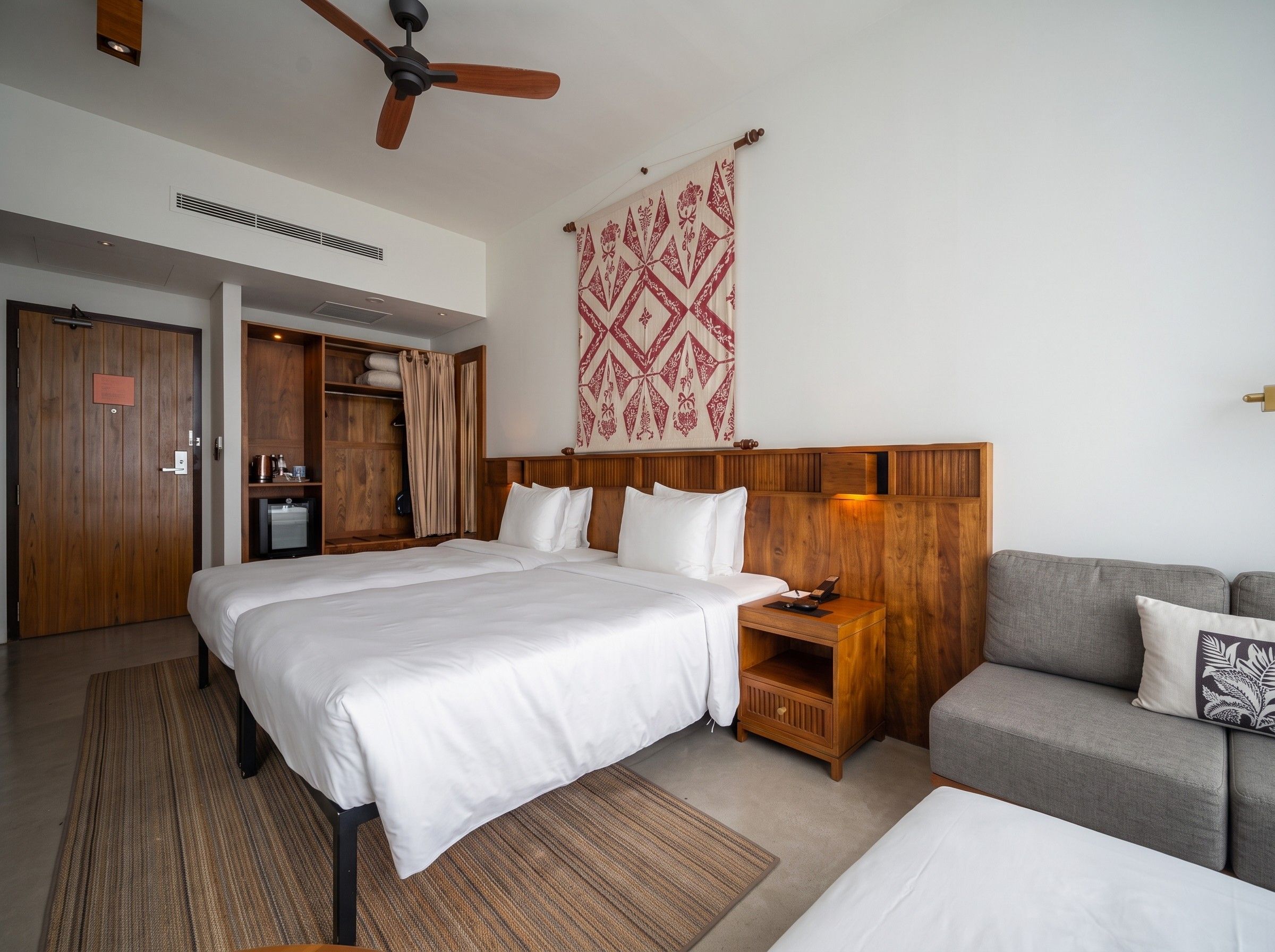 A hotel room with a large bed, a wooden headboard, a grey sofa, a small table, and a ceiling fan.