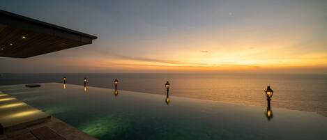Ein Fernblick auf einen Sonnenuntergang von einem Infinity-Pool, mit überdachtem Bereich und drei beleuchteten Laternen.
