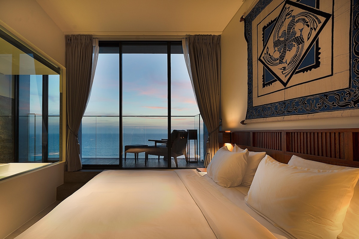Une chambre d’hôtel avec un grand lit, une vue sur la mer, un balcon avec une table et des chaises, et un élément décoratif mural.