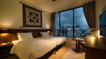 Habitación de hotel con dos camas, un escritorio, una silla, un ventanal amplio con vista a la ciudad y un adorno decorativo en la pared.