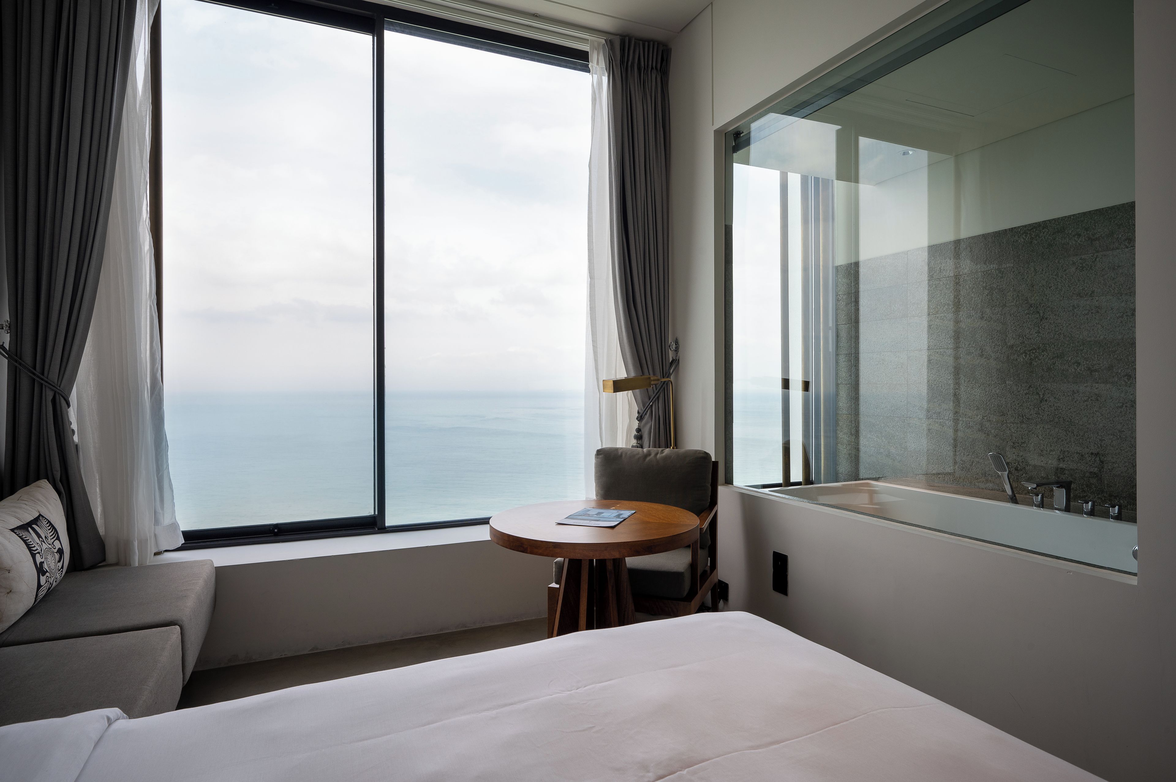 A hotel room with a bed, a small table, a chair, and a large window offering a view of the sea.