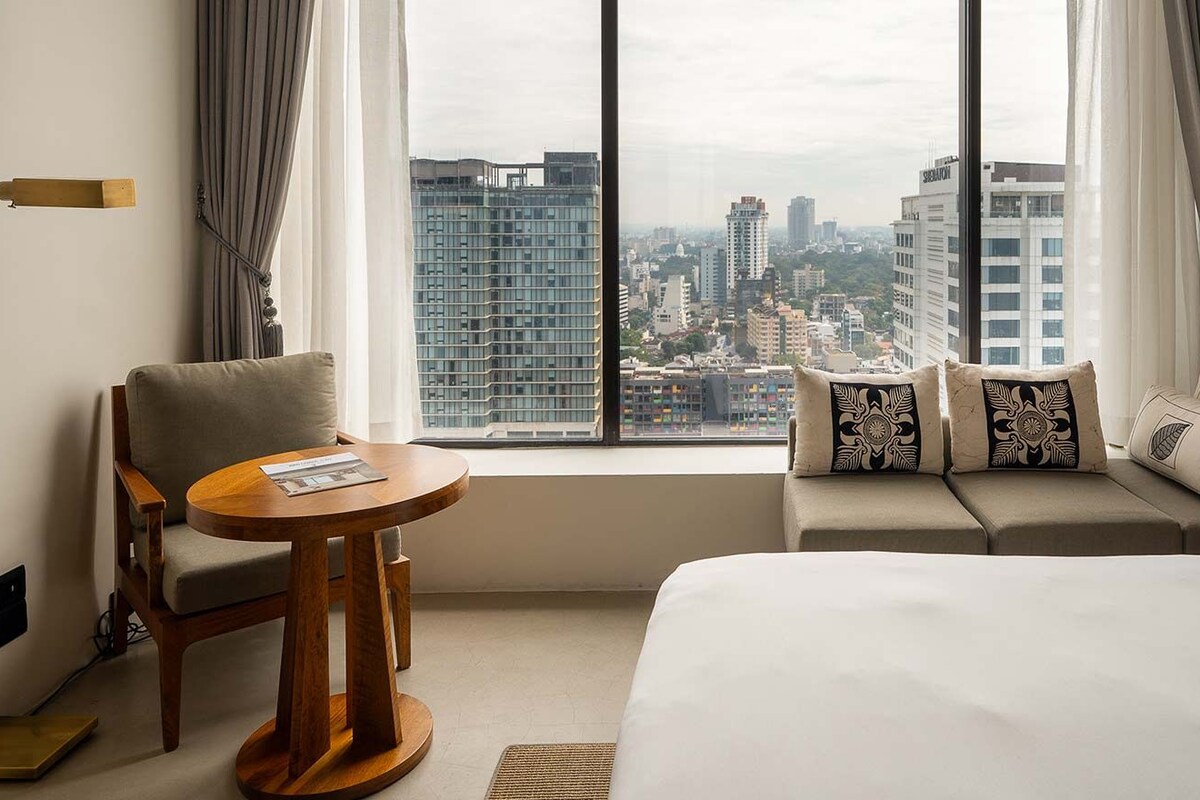 Habitación de hotel con una cama, un sofá con cojines estampados, una mesita redonda pequeña, una silla y vistas de un paisaje urbano a través de ventanas amplias.