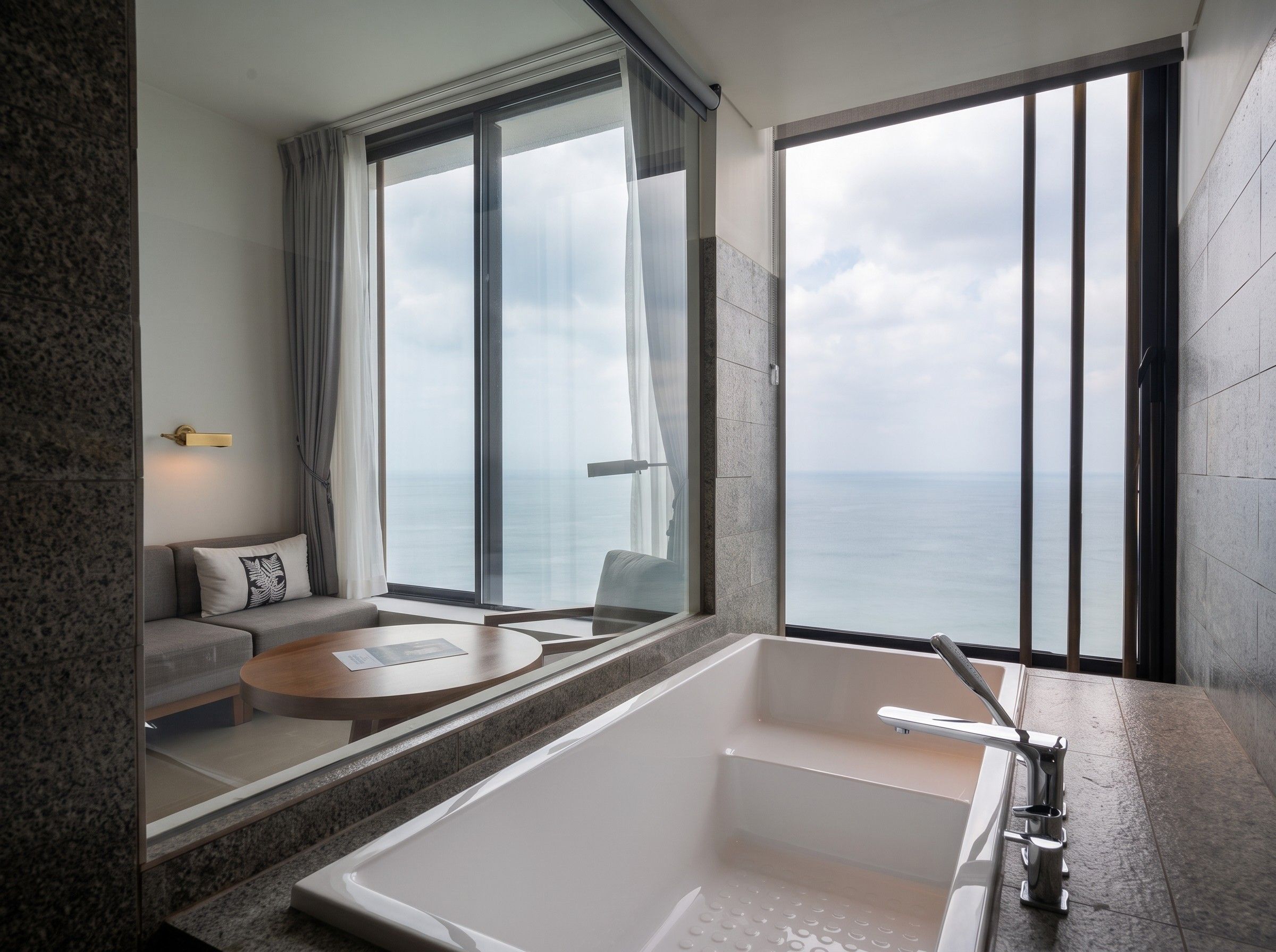Un baño moderno con una bañera grande, una zona de estar con sofá y mesa, y vistas al mar a través de amplios ventanales.