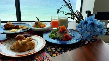 Una mesa con alimentos para el desayuno, incluyendo cruasanes, huevos y fruta, con vista al mar.