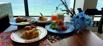 Una mesa con alimentos para el desayuno, incluyendo cruasanes, huevos y fruta, con vista al mar.