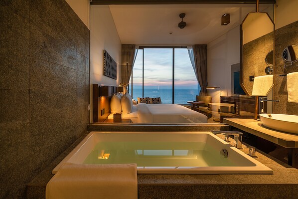 Una habitación de hotel moderna con una cama grande, una bañera, un ventanal con vistas al mar y un baño con lavabo y espejo.