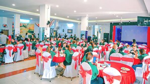 A conference room with round tables, white chairs with red sashes, and attendees in green shirts. - Ngoc Han Hotel By The Moment (Ho Chi Minh City)