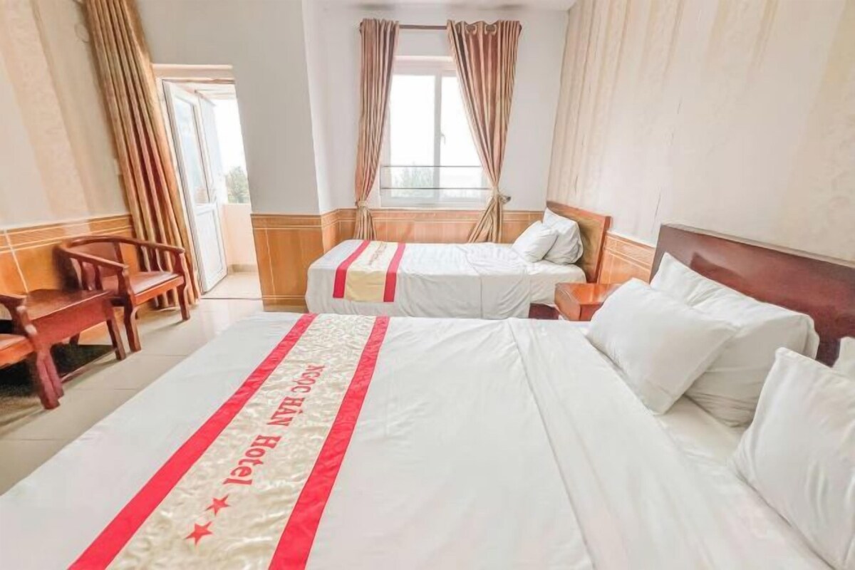 A hotel room with two beds, a wooden bench, and a view of the outdoors through a window with curtains.