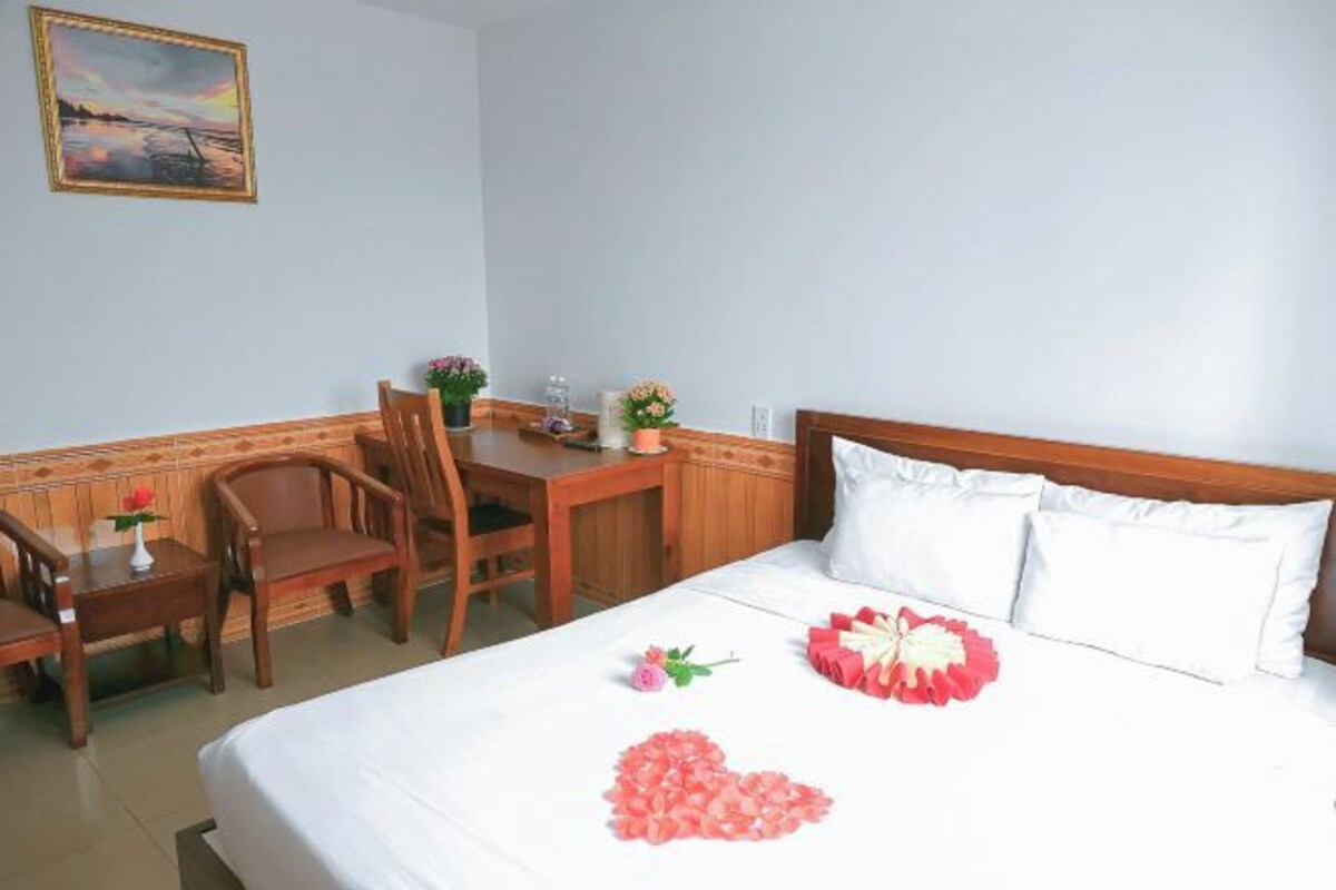 Une chambre d’hôtel avec un lit, un bureau en bois, une chaise et une petite table sur laquelle est posé un vase de fleurs.