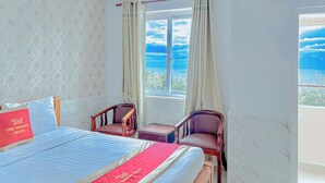 Ein Hotelzimmer mit einem Bett, zwei Stühlen, einem fensterbelaubten Fenster und Blick aufs Meer.