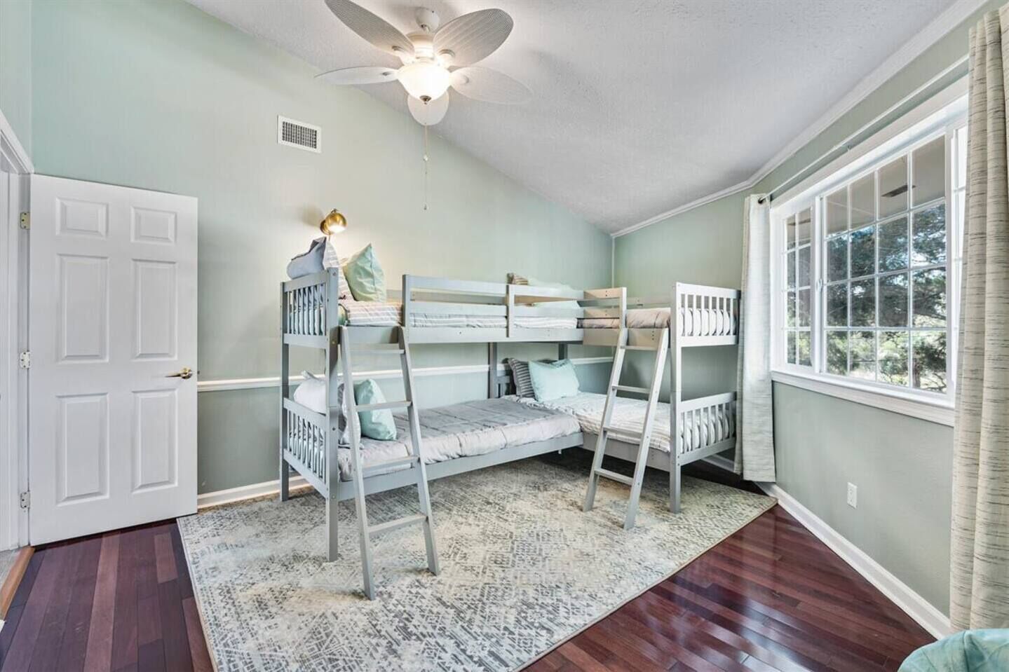 A room with a bunk bed, a ceiling fan, a window with curtains, and a door.