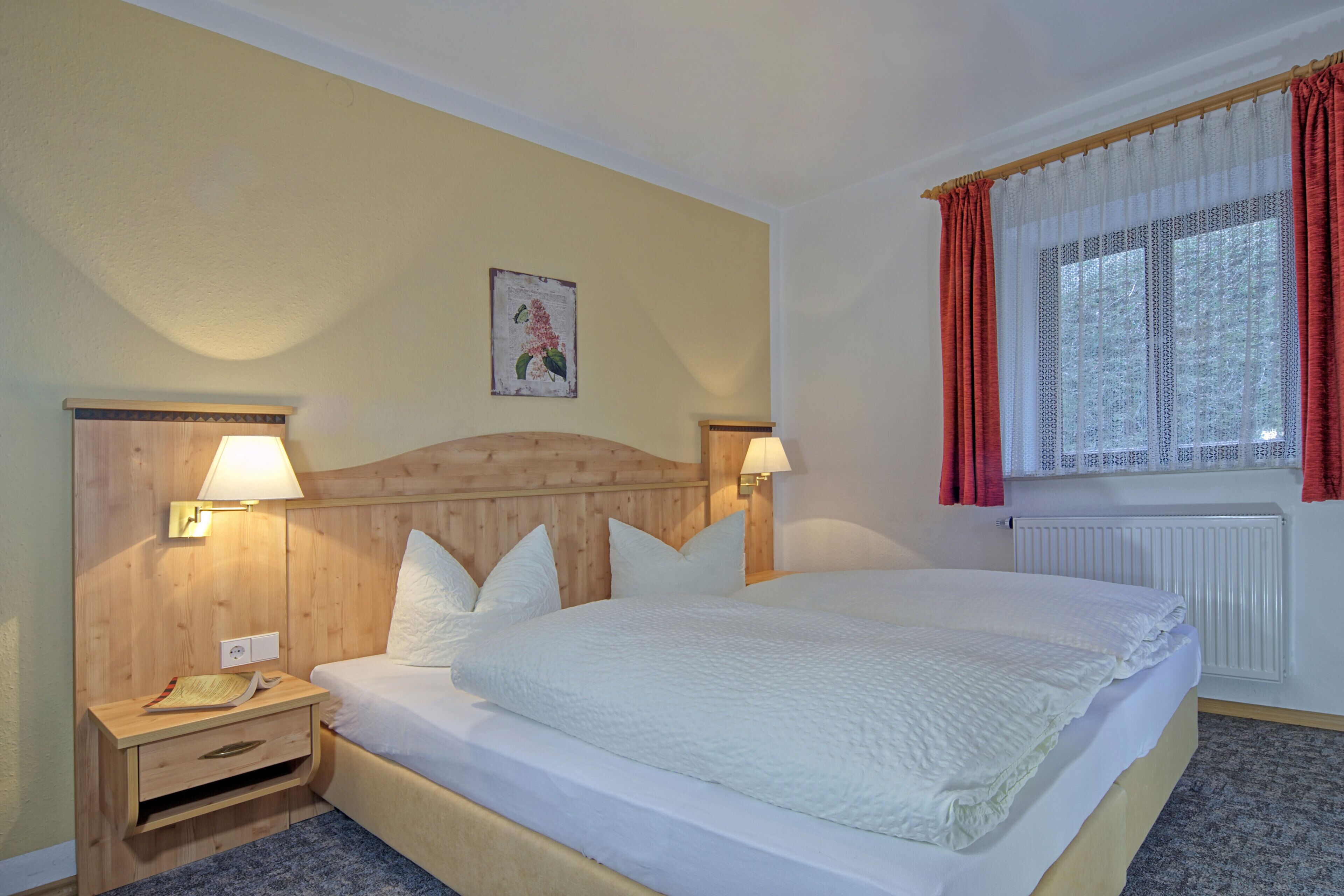 A hotel room with a wooden headboard, two bedside lamps, a large bed with white bedding, a small bedside table with a lamp, and a window with red curtains.