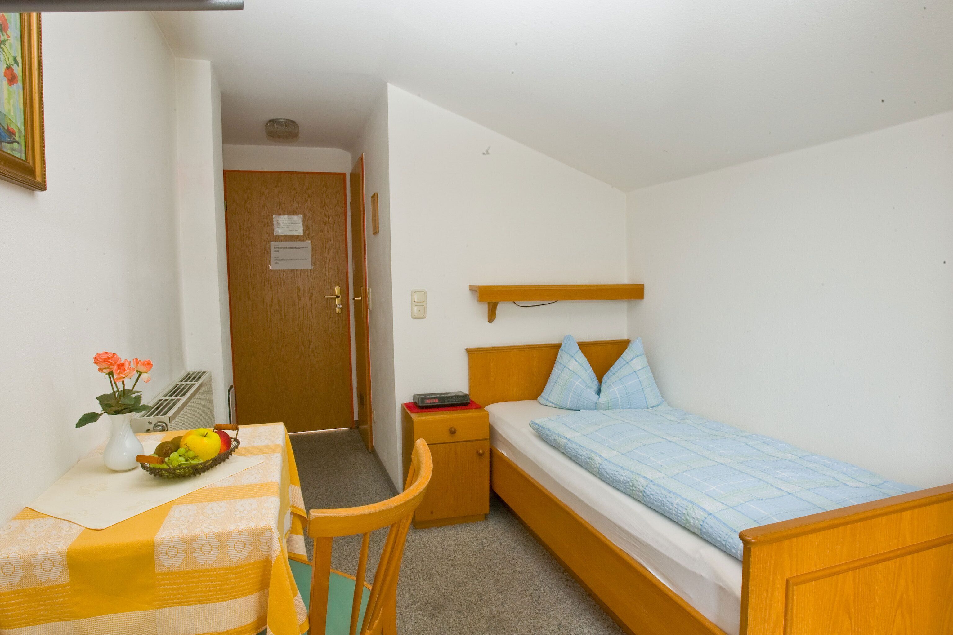 A small, single-room accommodation with a bed, a bedside table, a chair, a small table with a vase of flowers, and a wall-mounted shelf.