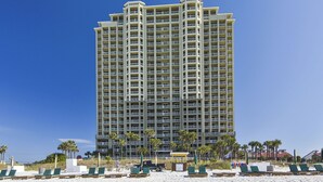 A tall hotel with multiple balconies, a sandy beach in the foreground, and palm trees.