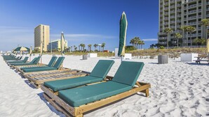 A beach with rows of wooden lounge chairs and green cushions, a tall hotel building, and palm trees in the background.