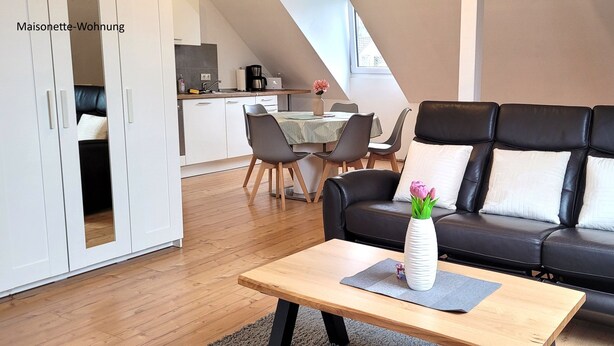 A modern living room with a black leather sofa, a wooden coffee table, and a dining area with a table and chairs. - Vacation home Südwind - Maisonette apartment no. 2 (Rhauderfehn)