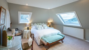 Un dormitorio con una cama grande, dos mesitas de noche con lámparas, una pequeña mesa con un espejo y flores, y dos tragaluces.