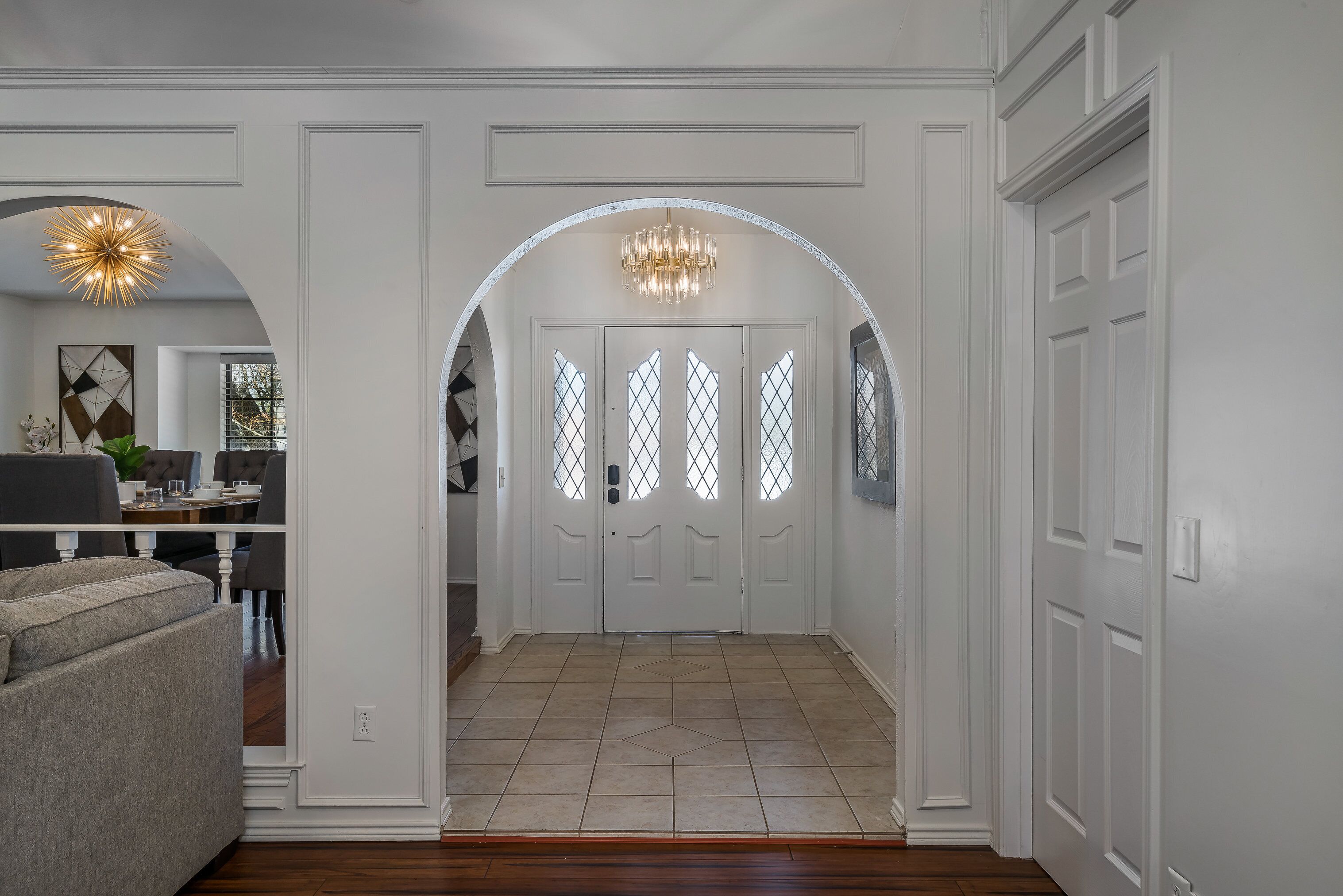 A hallway with a white door, a chandelier, and a dining area visible through the door.