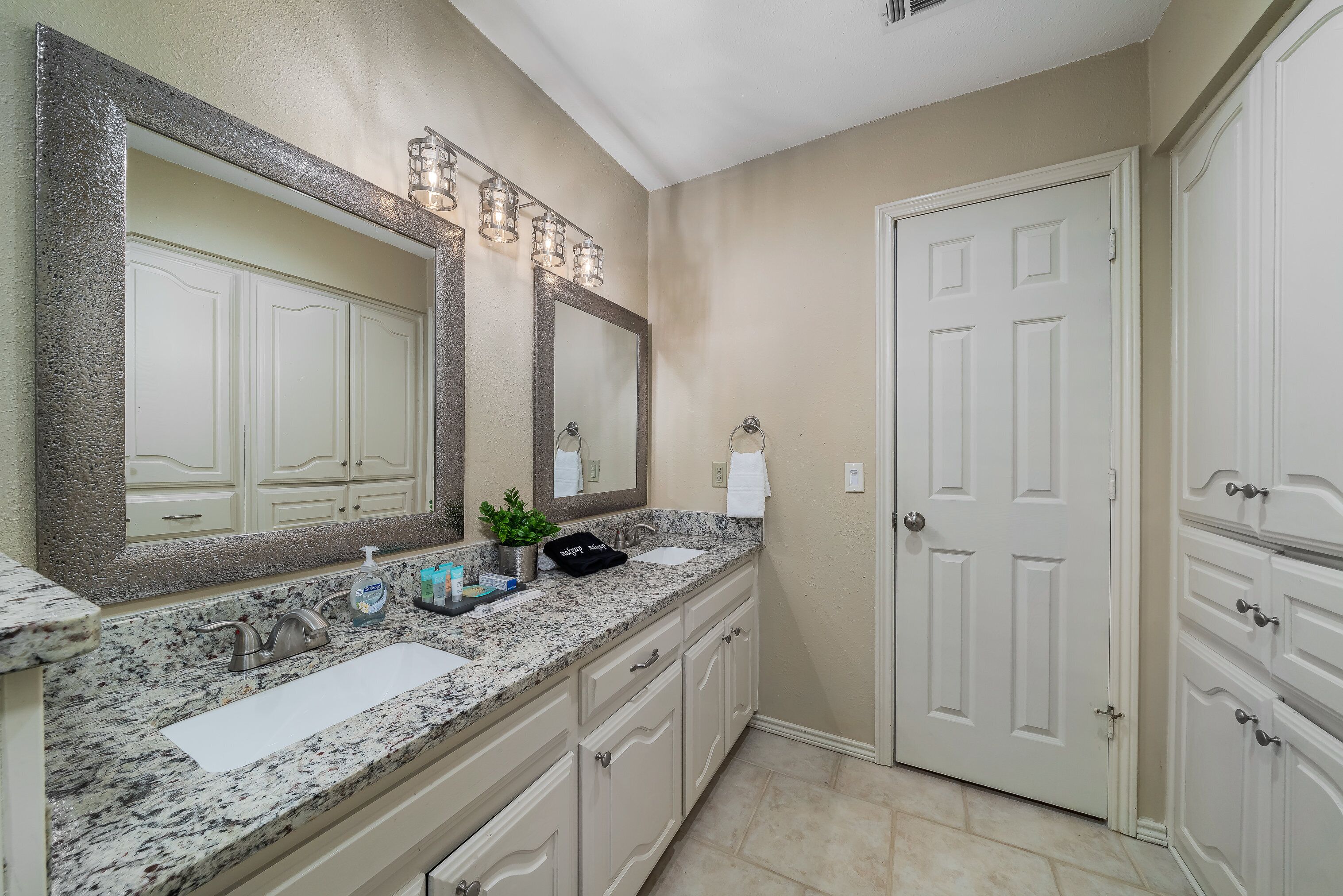A bathroom with a granite countertop, two sinks, a large mirror, and a door.