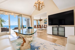 A spacious living room with a large glass table, a television, and a view of the ocean.