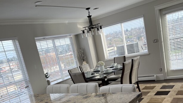 Una cocina moderna con encimeras de granito, un comedor con mesa de vidrio y ventanas grandes con persianas.
