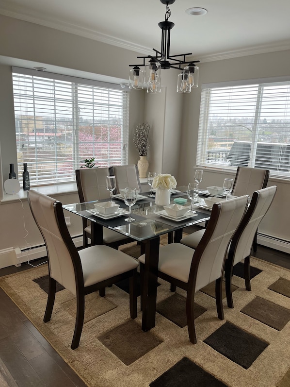 Une salle à manger avec une table en verre, six chaises, un lustre et de grandes fenêtres équipées de stores.