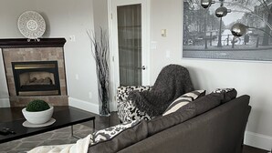 Un salón con un sofá gris oscuro, un plaid blanco, una alfombra estampada, una chimenea y un cuadro de un paisaje urbano en la pared.
