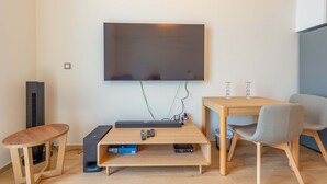 Una sala de estar moderna con un televisor de pantalla plana montado en la pared, una mesa de centro de madera con dispositivos electrónicos, una mesita auxiliar y un sillón gris.