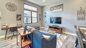 A modern living room with a large window, a flat-screen TV, a wooden desk, a blue sofa, and a clock on the wall. - Salida Hideaway- New Studio Condo (Salida)