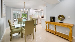 A modern dining area with a glass table and chairs, a yellow console table, and a view of the kitchen and outdoor area. - ‘Pelican Nest’, Tropical Naples Villa w/ Pool (Naples)