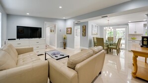 A modern living room with a flat-screen TV, beige sofas, a dining area with a glass table, and a kitchen with white cabinets. - ‘Pelican Nest’, Tropical Naples Villa w/ Pool (Naples)