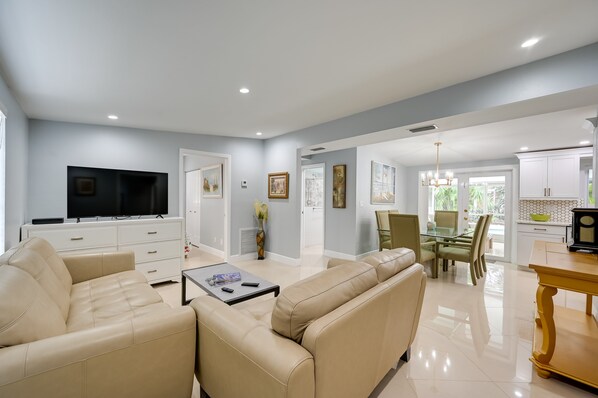 A modern living room with a flat-screen TV, beige sofas, a dining area with a glass table, and a kitchen with white cabinets.